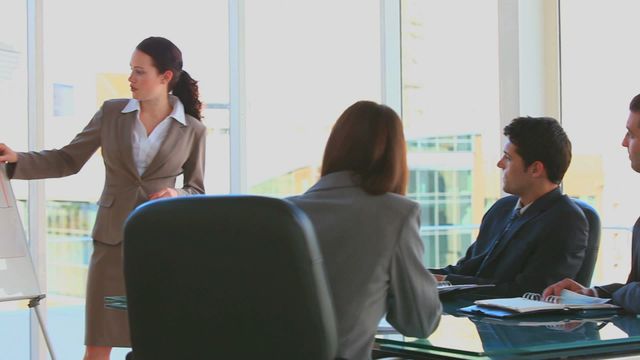 Businesswoman is presenting to team, pointing at flip chart in modern office. Coworkers are attentively listening while seated at glass table. This can be used for illustrating teamwork, professional environment, office meetings or corporate training.