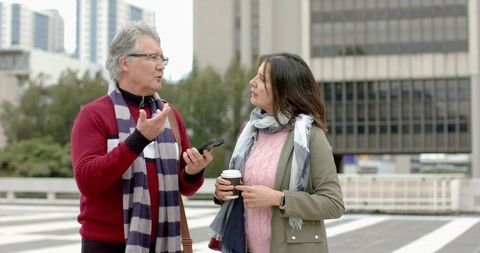 Mature couple conversing in urban plaza holding smartphone and coffee, casual winter attire