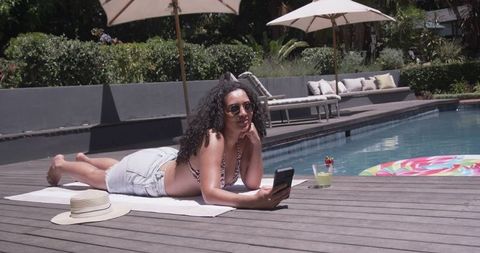African american woman lounging poolside, sunbathing and browsing smartphone with cocktail