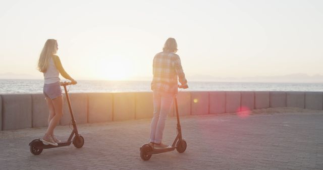 Young couple enjoying a leisurely ride on electric scooters by the beach at sunset. They are riding on a paved path near the ocean, with the sun low on the horizon, creating a serene and beautiful setting. Ideal for promoting outdoor activities, romantic getaways, eco-friendly transportation, or coastal lifestyle imagery.