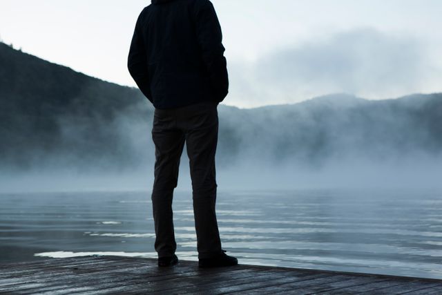 Man is seen standing on deck, observing fog over a lake, ideal for themes of solitude, tranquility, introspection, or travel. Suitable for blog posts about nature retreats, personal growth, or peaceful getaways.