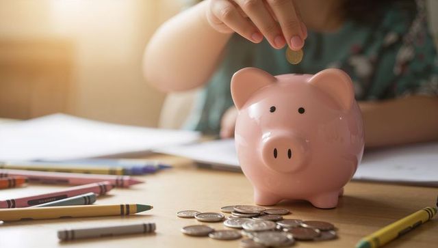 Child dropping coin into pink piggy bank, saving money with coins and crayons on table