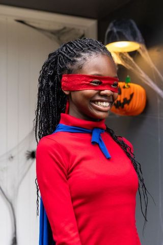 Smiling african american girl wearing red superhero costume at front door with pumpkin
