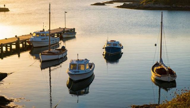 Small boats moored in golden-hour harbor reflecting on calm cove water by wooden pier