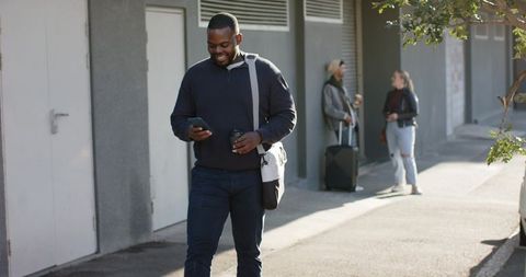 Young African American Man Walking Urban Sidewalk Checking Phone Holding Coffee and Shoulder Bag