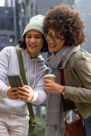 Two friends laughing and checking smartphone while walking urban street with coffee and knitwear