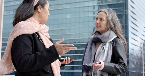 Mature women colleagues discussing outdoors near glass office building wearing scarves