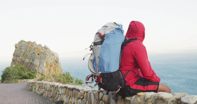 Hiker with large backpack sitting on rocky ledge overlooking coastline. Perfect for adventure and travel marketing, outdoor activity blogs, nature exploration articles.