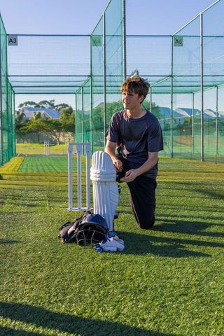 Teenage cricketer kneeling preparing pads helmet gloves stumps at outdoor practice nets