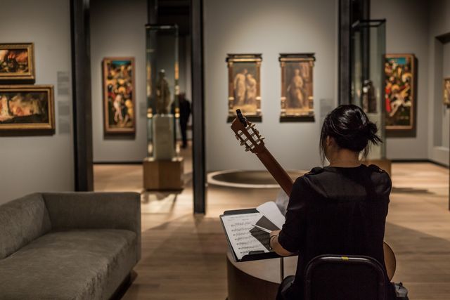 This image depicts a musician practicing classical guitar in an art gallery. She is seated with a guitar, looking at sheet music, with various paintings visible on the walls around her. This image can be used to represent the intersection of music and visual art, cultural events, art exhibitions, and the serenity of artistic practice within historical settings. Ideal for materials related to music performances, art gallery promotions, or cultural appreciation campaigns.