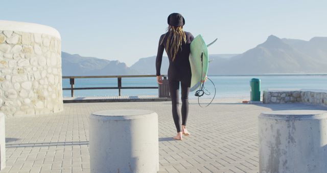 Woman carrying green surfboard walking on beachside path. Mountains and ocean in background. Ideal for surfing, outdoor lifestyle, summer activities, and travel promotions.