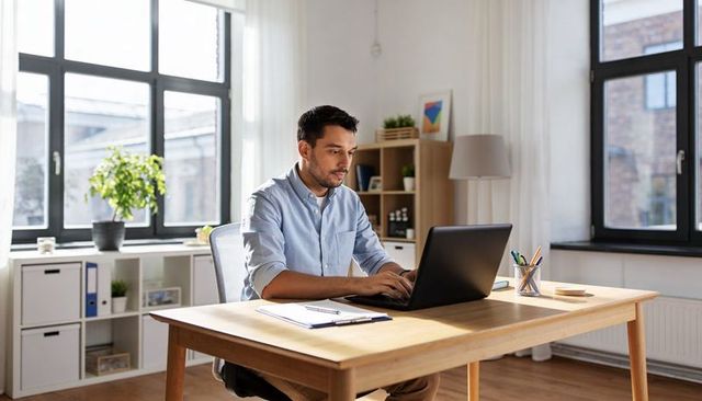 Man working at minimalist sunlit home office with laptop, plant and organized desk space