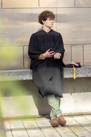 Campus graduate leaning on wall holding smartphone beside mortarboard tassel and diploma