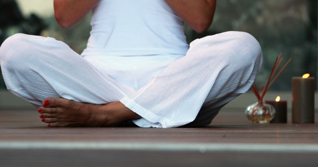 Person sitting cross-legged on floor, wearing white clothing. Candlesticks and incense holder in background. Ideal for yoga and meditation environments, wellness retreats, or mindfulness practices. Conveys tranquility and focus.