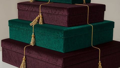 Stacking jewel-tone velvet gift boxes with gold braided tassels on neutral tabletop