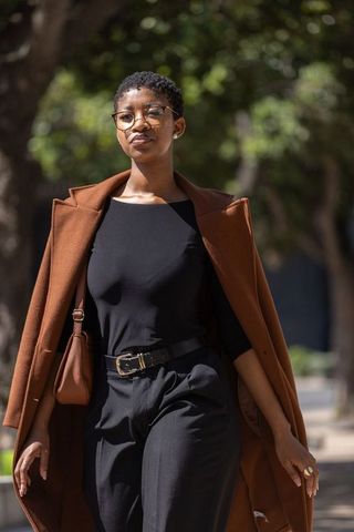 Confident woman walking in rust coat and round glasses with brown satchel on city street