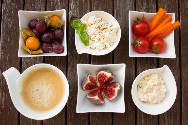 Arrangement of various healthy breakfast items on wooden table. Set includes fruits like grapes and figs, vegetables like tomatoes and carrot sticks, cottage cheese with spices, cream dip, and cup of coffee. Ideal for health-focused food blogs, nutrition articles, or culinary presentations.