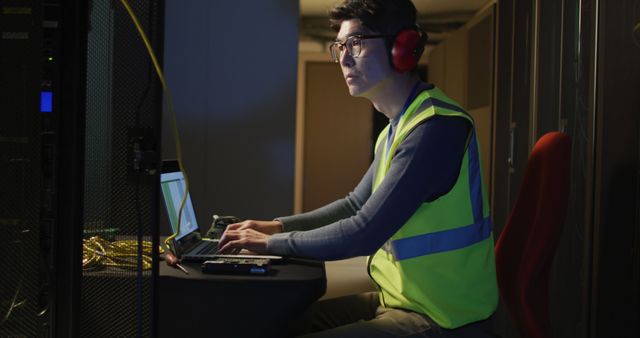 A technician wearing a high visibility vest and ear protection monitoring network equipment in a data center. He is seated at a desk, working on a laptop, focusing intently on the screen. This image can be used in articles, blogs, or advertisements related to information technology, data centers, network management, or cybersecurity.