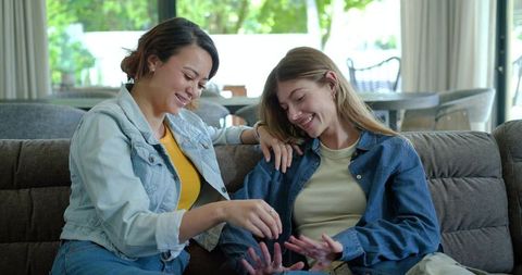 Two women sharing playful at-home manicure on sofa, cozy denim jackets, natural daylight
