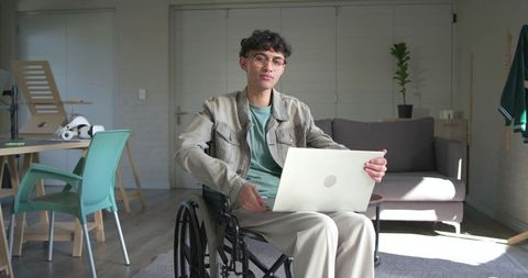 Young asian man in wheelchair working on laptop in sunlit minimalist home office