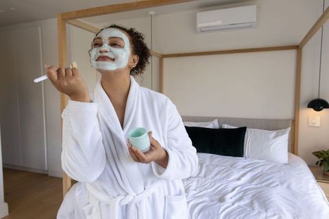 African american woman applying facial mask in robe in modern bedroom, morning self-care