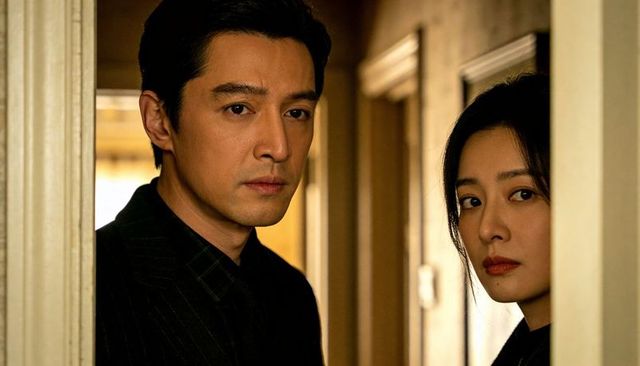 Middle-aged couple standing in hallway wearing dark formal outfits, intense expressions