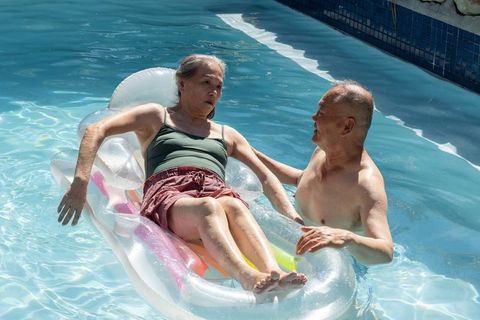 Diverse senior couple enjoying pool float with man assisting woman on inflatable raft