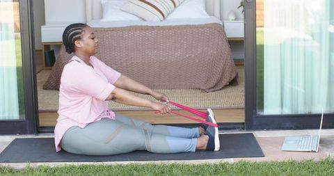 African american woman doing seated resistance band stretch on patio doorway