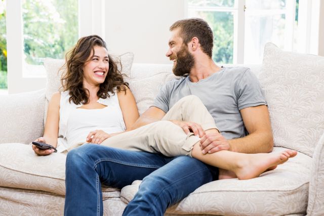 Couple sitting on a comfortable sofa in a bright living room, smiling and enjoying each other's company. Ideal for use in advertisements, blogs, or articles about relationships, home life, and leisure activities. Perfect for illustrating themes of love, happiness, and domestic comfort.
