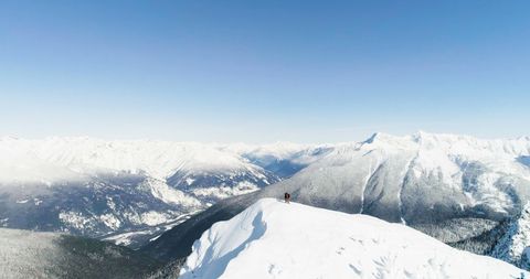 Person standing triumphantly on snow-covered mountain peak with breathtaking panoramic views of surrounding landscape. Ideal for outdoor adventure promotions, winter travel brochures, hiking and winter sport advertisements, inspirational and motivational posters.