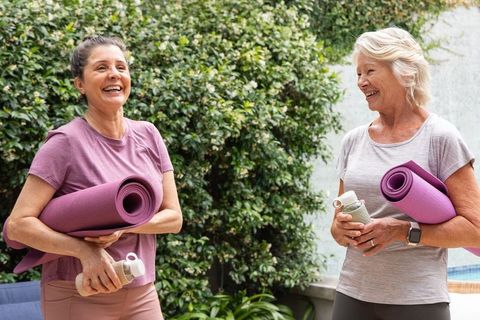 Senior women laughing while holding purple yoga mats and water bottles poolside in garden