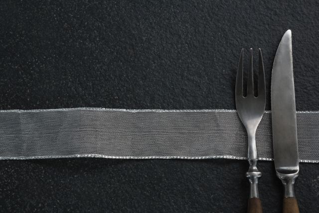 Elegant fork and knife placed on a black slate background with a silver ribbon. Ideal for use in restaurant menus, culinary blogs, kitchen decor, and dining-related advertisements.