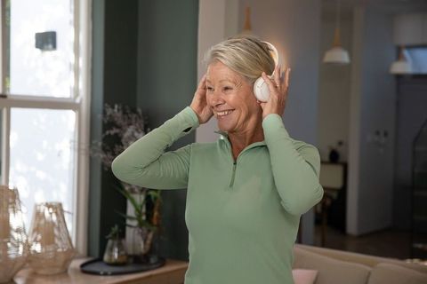 Senior woman smiling and adjusting wireless headphones in sunlit cozy living room