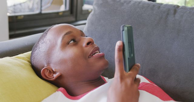 Young boy holding smartphone and using voice command while relaxing on couch. Captures contemporary moment with technology and leisure. Ideal for technology, family lifestyle, and communication themes.