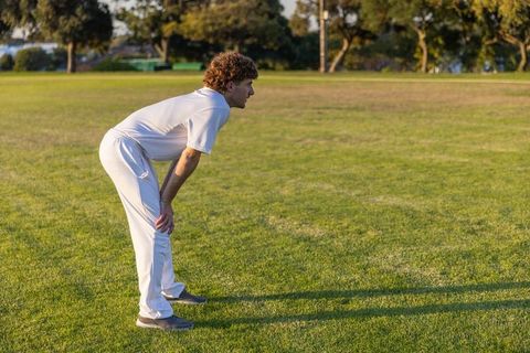 Cricket fielder bending hands-on-knees on sunlit grass during outdoor training session