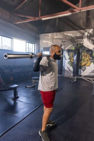 Bearded man carrying barbell on shoulders in industrial gym with mural and punching bags