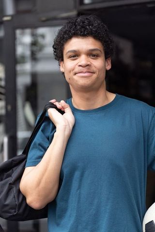 African american young man holding soccer ball and backpack smiling urban casual portrait