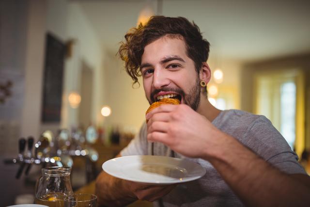 Man enjoying a croissant in a cozy café, smiling and looking content. Ideal for use in lifestyle blogs, food and beverage promotions, café advertisements, and social media content highlighting casual dining experiences.