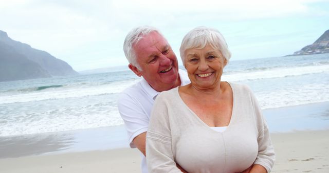 A senior Caucasian couple shares a joyful moment on a beach, with copy space. Their smiles and closeness suggest a deep bond and contentment in each other's company.