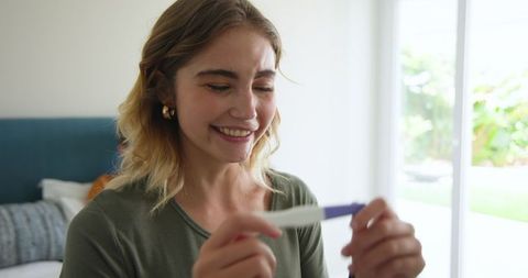 Joyful woman holding positive pregnancy test in bright bedroom