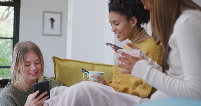 Happy diverse teenager girls sitting on sofa, drinking cocoa and using smartphones. Spending quality time, lifestyle, friendship and adolescence concept.