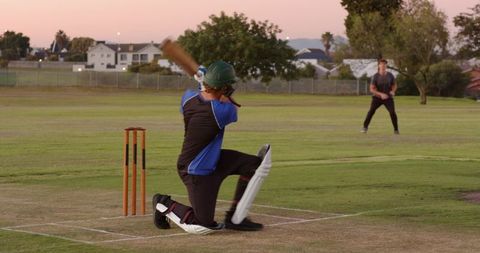 Kneeling cricket batter hitting powerful shot during twilight match with distant fielder
