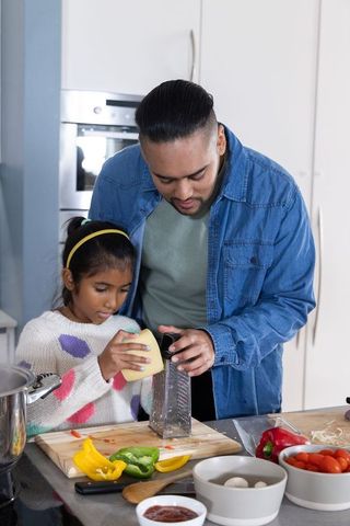 Indian father guiding daughter grating cheese at kitchen counter for family cooking
