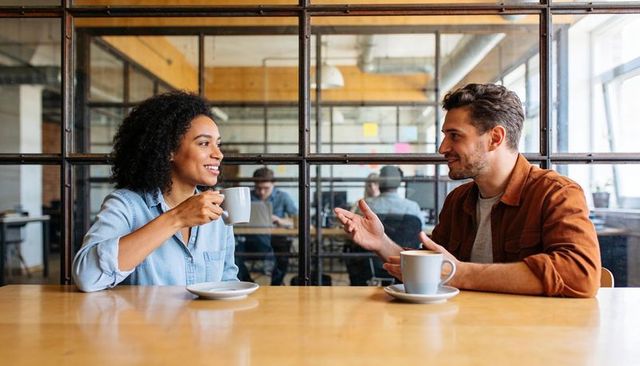 Coworkers sharing coffee and conversation at modern office table by glass partition