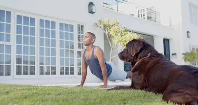 Focused biracial man practicing yoga with dog lying in sunny garden. Summer, wellbeing, fitness, pet and healthy lifestyle concept, unaltered