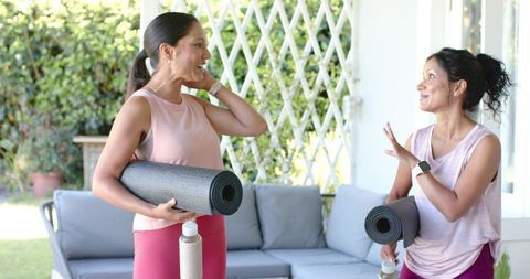 Multicultural women chatting on patio holding yoga mats and water bottles in activewear