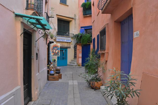 Charmingly narrow European alley with vibrant pastel buildings adorned with potted plants provides a picturesque and inviting atmosphere. Ideal for travel blogs, architectural features, or urban lifestyle content focusing on serene and charming village settings.