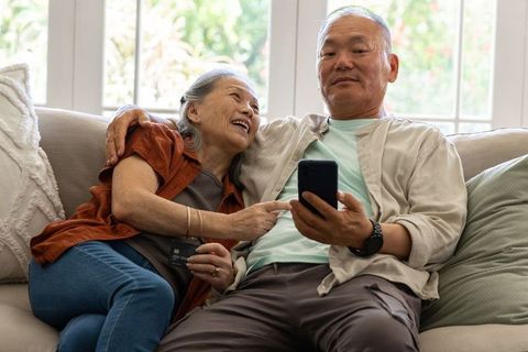 Senior asian couple relaxing on sofa sharing smartphone and credit card, cozy home moment