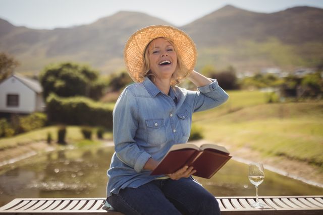 Senior woman enjoying a sunny day outdoors while reading a book on a park bench. She is smiling and wearing a hat, with a wine glass beside her. Ideal for use in lifestyle, leisure, and senior living content, promoting relaxation and happiness in nature.