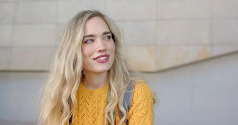 Smiling blonde woman standing and looking away in mustard sweater with backpack in city daylight